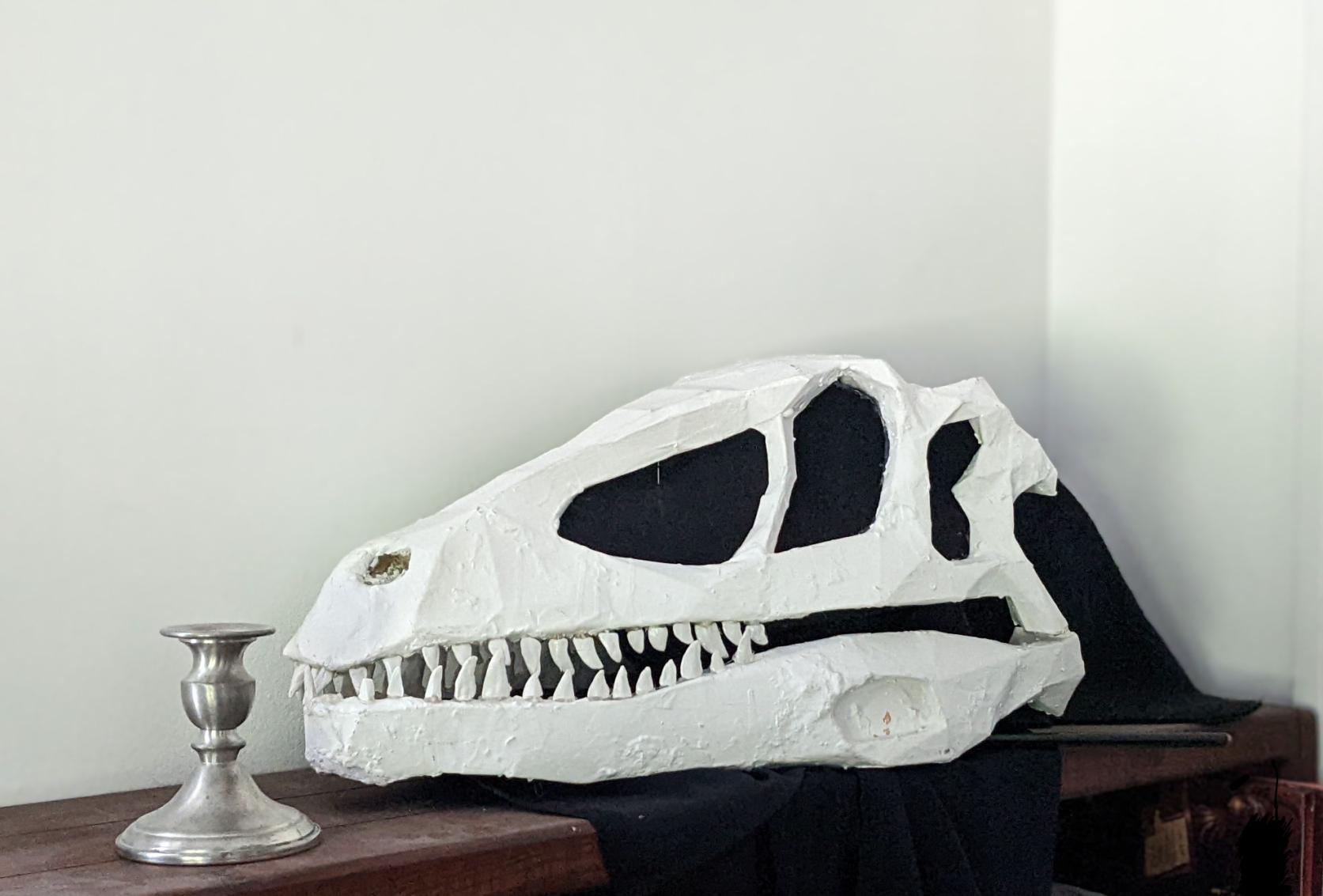 A white dino skull rests on a dark wood sideboard, with a silver candlestick at one end. The skull drips black fabric, which pools under the skull before running off the sideboard