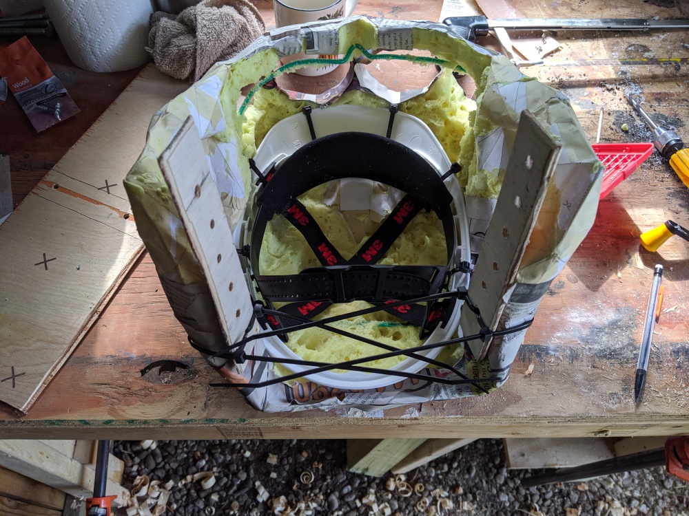 The skull sits on a workbench. The zipties threaded through the wire armature now secure the brim of a construction helmet and its liner to the inside of the skull, and two wooden posts are anchored at the rear of the skull with several holes drilled through them.