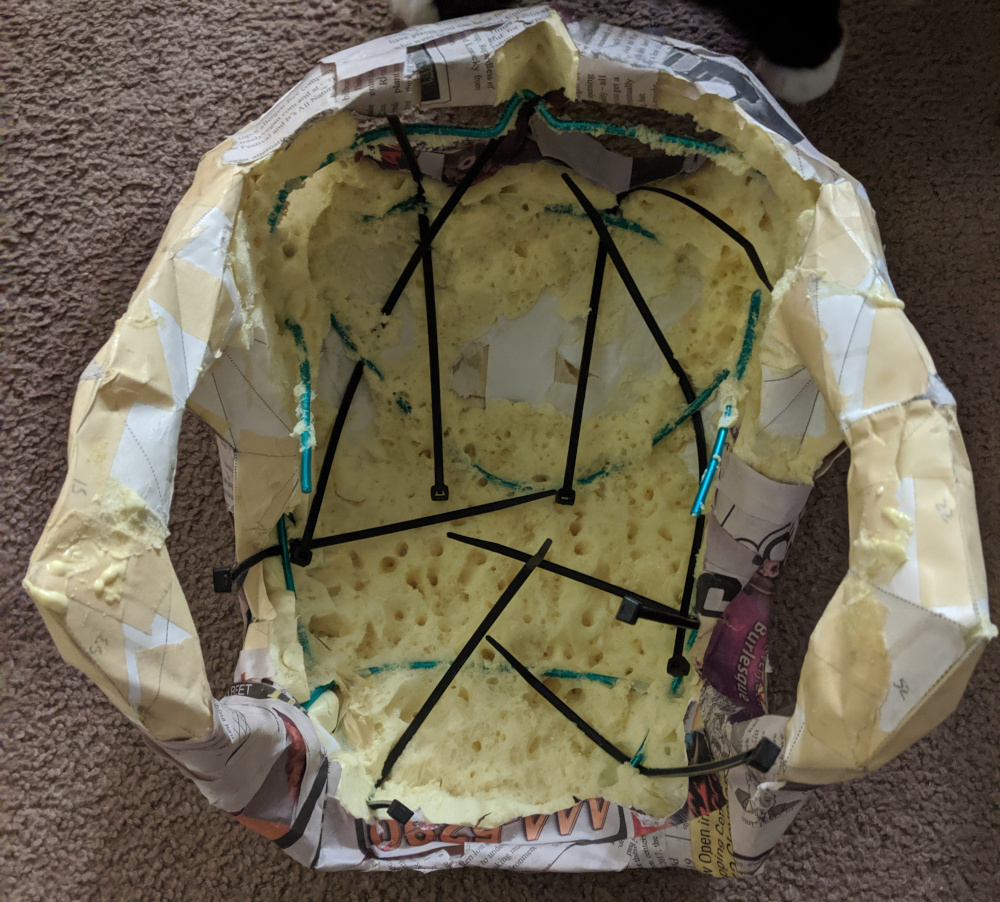 Sitting on brown carpet, the inside of the skull is shown. The formerly-visible bulges of foam have been removed, leaving green wires nestled in a see of yellow froth. Black zipties are threaded through the wires, but not yet zipped.