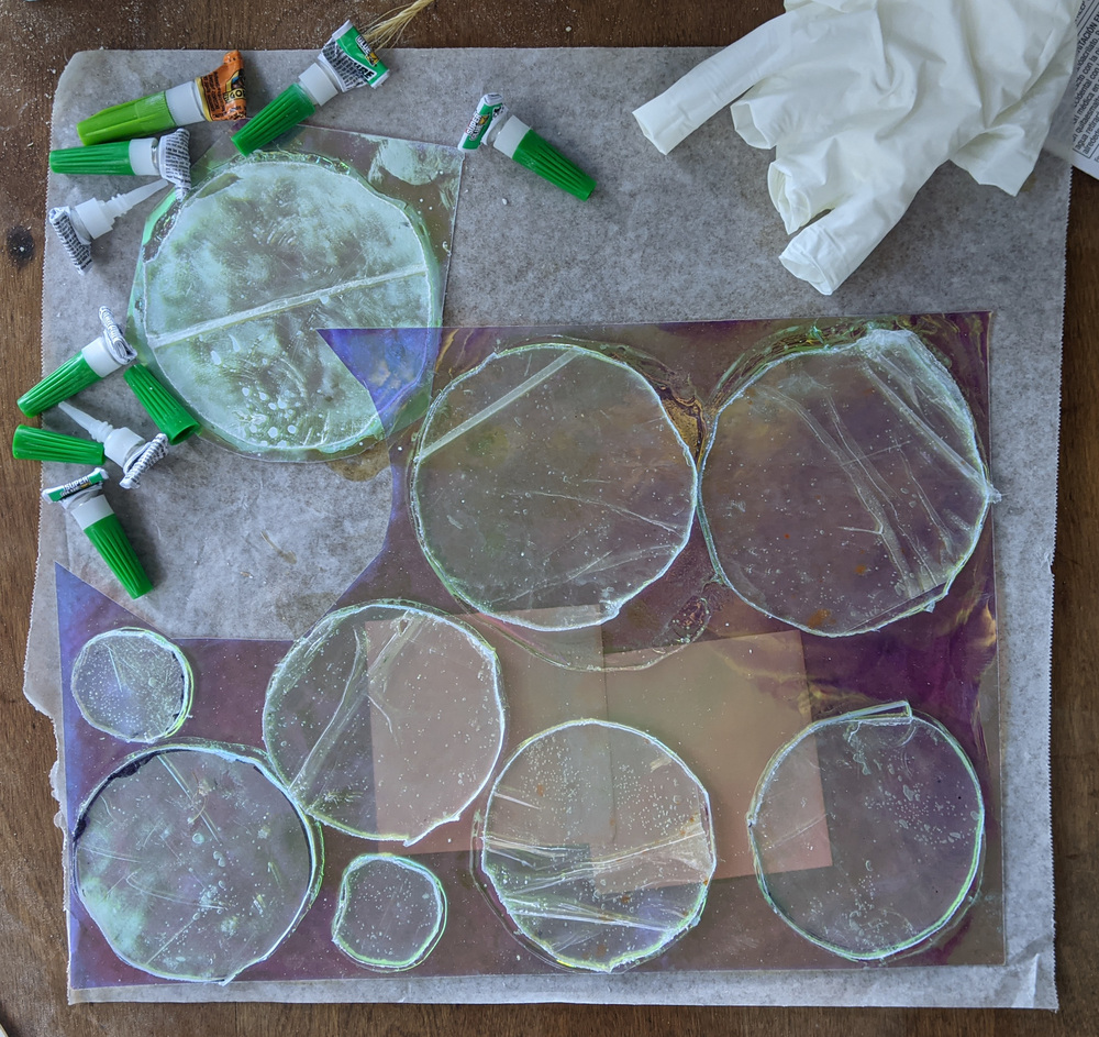 On a wooden tabletop, on a sheet of waxed paper, a sheet of metallic acrylic film is laid, covered in round plexiglass disks which have been glued down. Eight empty superglue tubes are piled in a corner, in another corner a discarded nitrile glove. One disk has been cut out of the acrylic sheet.