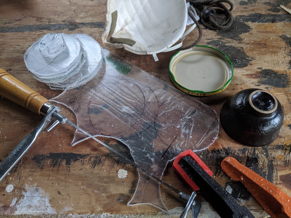 On a wooden workbench. Arranged artfully: a sheet of plexiglass with some circles drawn on its protective cling film, and some circles of plexiglass cut from that sheet, the coping saw that cut them, some clamps, a small tea bowl, a medium salsa lid, a N95 face mask.