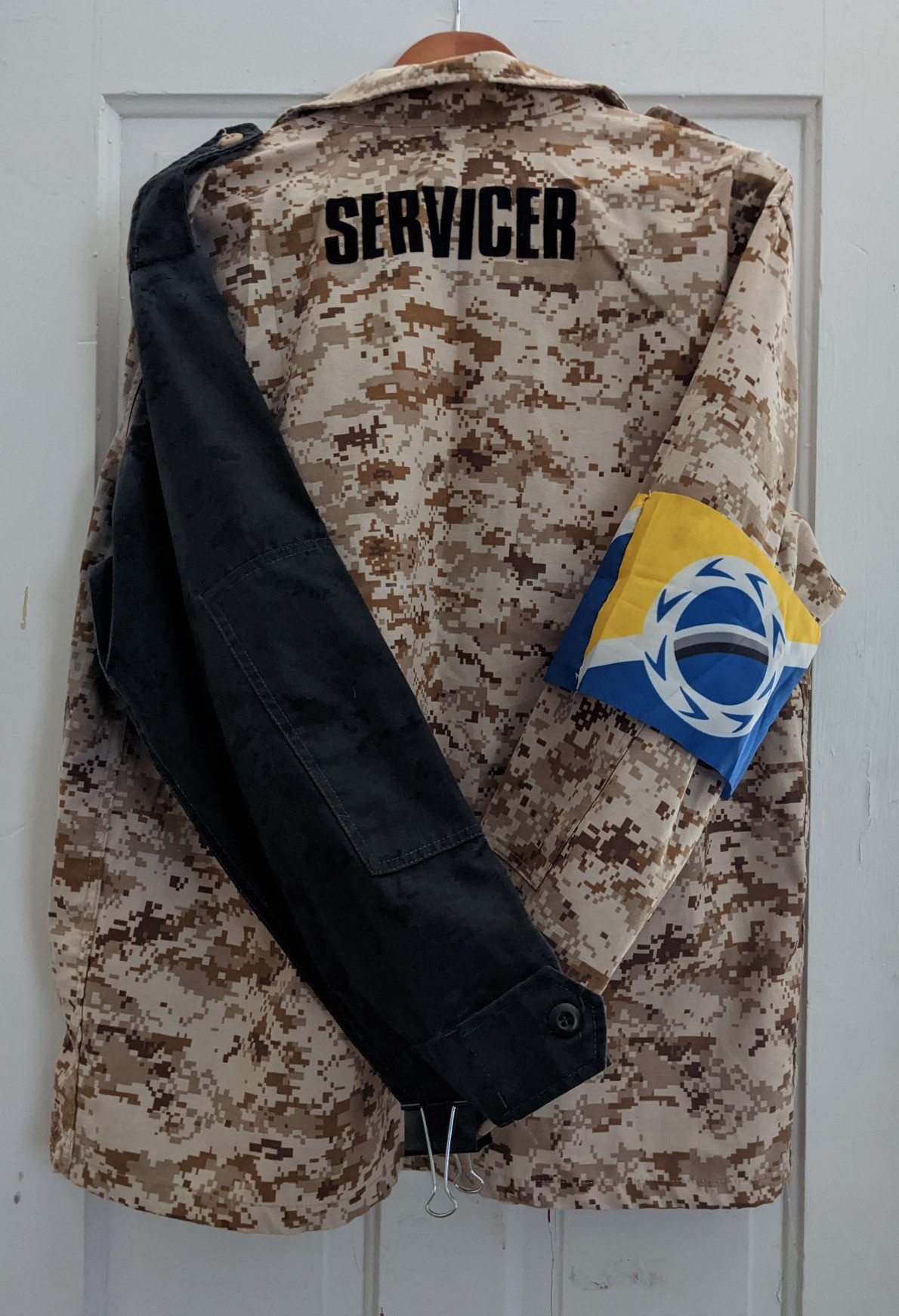 a desert camo coat hangs against a white wall. the left sleeve is dyed black; the right sleeve bears an armband made of yellow, white, and blue flags. Across the shoulders reads the word SERVICER in 1.5 inch high letters.
