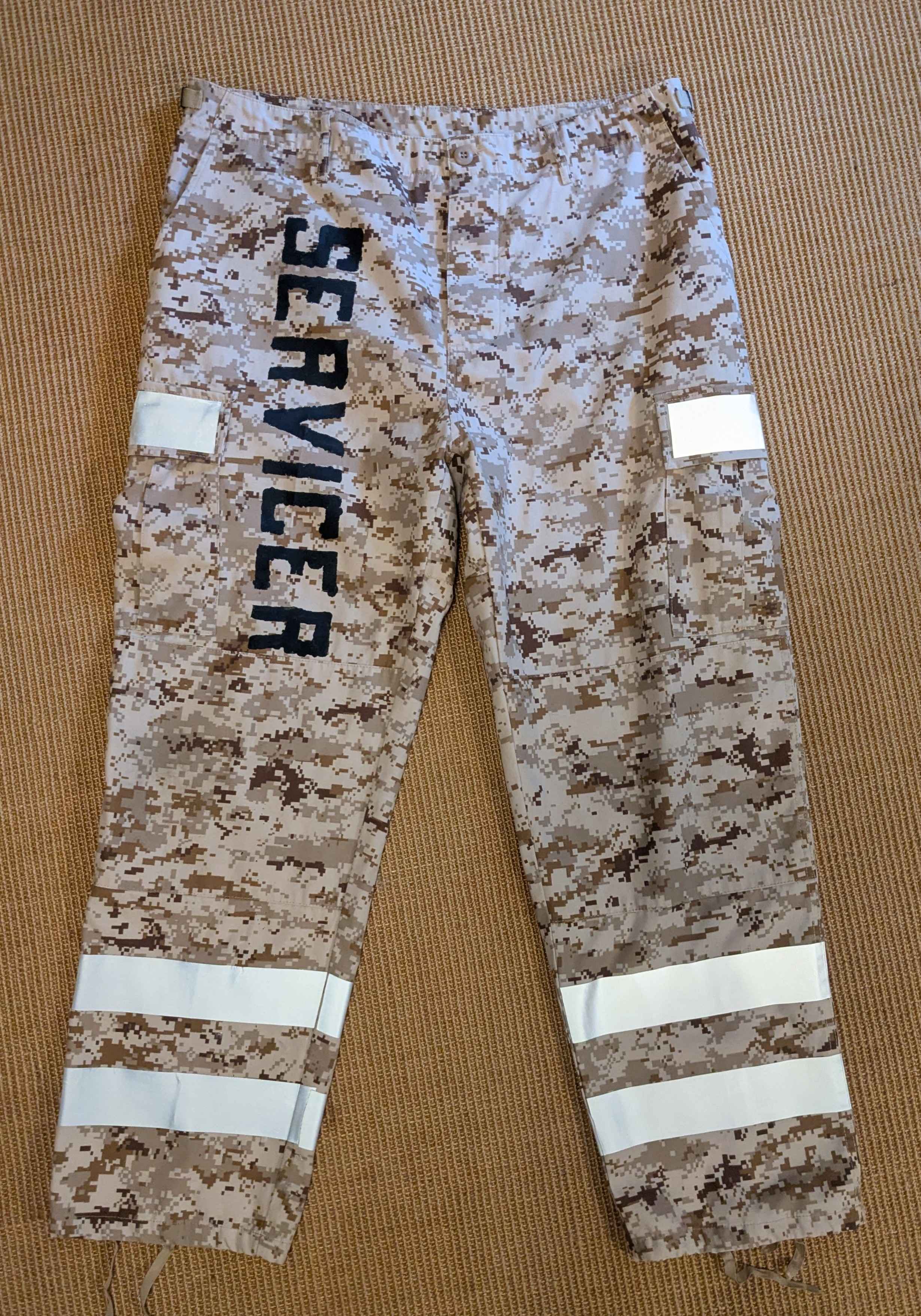 a pair of desert camo pants lie flat on the floor. retroreflective tape makes bands twice around the calbes, and covers the flaps of the thigh pockets. on one thigh is stenciled the word SERVICER.